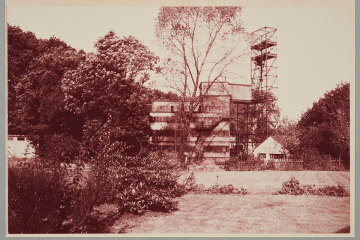 Hinter Gärten im Wald stehendes stählernes Gerüst und ein Gebäude sowie ein brückenartiges Gebilde