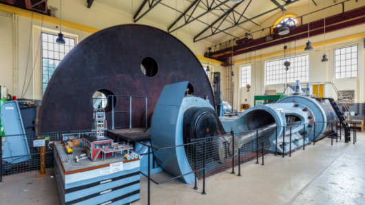 Eine Dampfmaschine mit einer großen Seiltrommel steht in einer Halle.